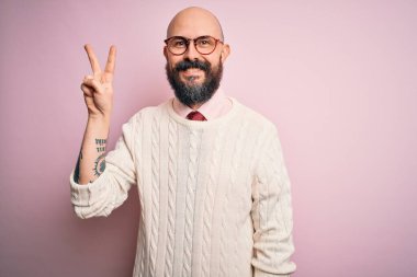 Sakallı, dövmeli, pembe arka planda gözlüklü ve süveterli yakışıklı bir adam. İki numaralı parmaklarıyla işaret ediyor. Kendinden emin ve mutlu bir şekilde gülümsüyor..