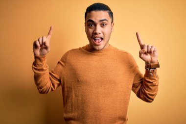 İzole edilmiş sarı arka planda dikilen sıradan bir süveter giyen genç Brezilyalı adam şaşkın şaşkın, parmaklarıyla işaret ediyor ve kollarını kaldırıyordu..