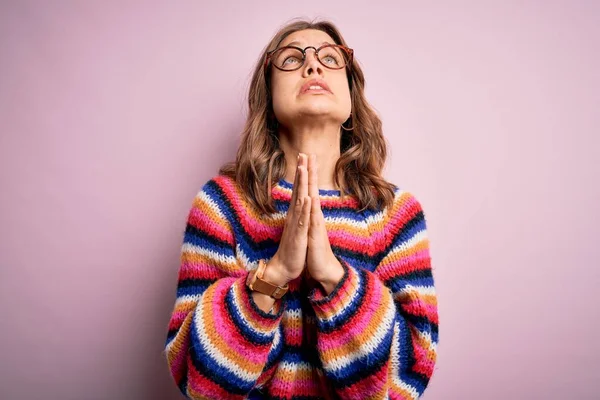 Young beautiful blonde girl wearing glasses and casual sweater over pink isolated background begging and praying with hands together with hope expression on face very emotional and worried. Begging.