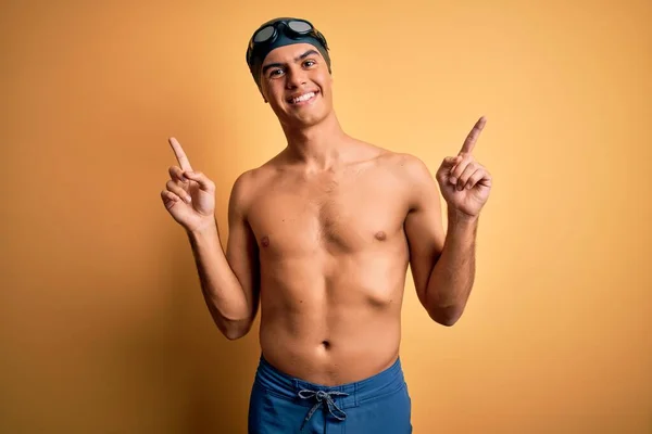 年轻英俊的男子穿着泳衣 戴着泳帽 披着孤零零的黄色背景 带着自信的微笑 手指指向不同的方向 广告的复制空间 — 图库照片