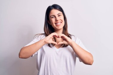 Genç, güzel, esmer, günlük tişört giyen, izole edilmiş beyaz arka planda duran, aşkla gülümseyen, elleriyle kalp sembolü yapan bir kadın. Romantik konsept.