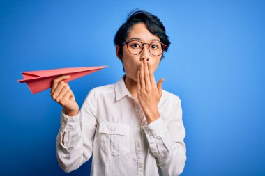 Genç, güzel Çinli bir kadın, elinde kağıt uçakla izole edilmiş mavi arka plan üzerini örtüyor. Ağzını kapatıyor, hata yaptığı için utanmış, korku ifadesi, sessizlikten korkmak, gizli bir konsept.
