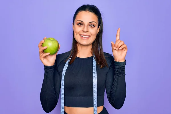 美しいブルネットスポーティな女性を使用してテープ測定幸せな顔を持つアイデアや質問指差し指で驚いて紫色の背景にリンゴの果実を食べる ナンバーワン — ストック写真
