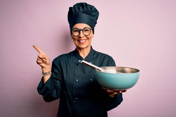 Middle age brunette chef woman wearing cooker uniform and hat using whisk and bowl very happy pointing with hand and finger to the side