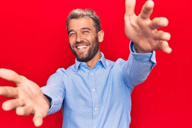 Genç, sarışın, gündelik tişört giyen, izole edilmiş kırmızı arka planda duran ve kucaklaşmak için kollarını açmış kameraya bakan yakışıklı bir adam. Mutluluğu kucaklayan neşeli bir ifade..