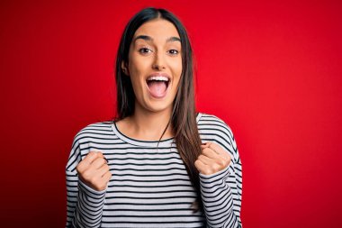 Genç, güzel, esmer bir kadın kırmızı arka plan üzerine sıradan çizgili tişört giyip kollarını kaldırarak ve açık gözlerle başarının şaşkınlığını kutluyor. Kazanan konsept.