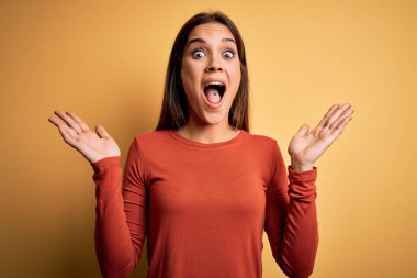 Sarı arka plan üzerinde sıradan tişörtler giyen genç ve güzel esmer kadın çılgın ve başarıyı kollarını kaldırıp heyecanla haykırarak kutluyor. Kazanan konsept