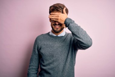 Sakallı, gözlüklü ve süveterli genç bir adam pembe arka planda durup gülümsüyor ve elleri yüzünde şaşkınlıktan gözleri kapayarak gülüyor. Kör kavram.