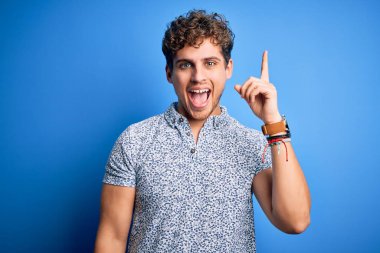 Genç, sarışın, tatilde, kıvırcık saçlı, polo giyen ve başarılı bir fikri işaret eden yakışıklı bir adam. Heyecanlı ve mutlu. Bir numara..