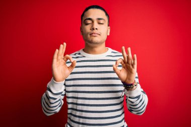 İzole edilmiş kırmızı arka planda dikilen sıradan çizgili tişört giyen genç Brezilyalı adam rahatlamış ve gözleri kapalı meditasyon hareketi yaparken parmaklarıyla gülümsüyor. Yoga konsepti.