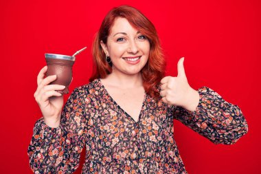 Genç, güzel, kızıl saçlı bir kadın izole edilmiş kırmızı arka planda içki içiyor mutlu ve pozitif gülümsüyor, başparmağıyla mükemmel ve onay işareti yapıyor.