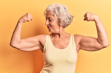 Üstü başı gri saçlı, sıradan kıyafetler giyen, gururla gülümseyen kol kaslarını gösteren yaşlı bir kadın. fitness konsepti. 