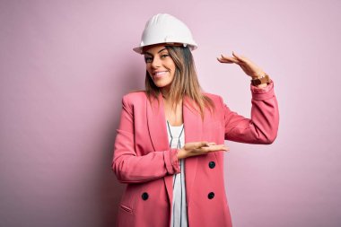 Genç ve güzel esmer mimar kadın pembe arka planda güvenlik kaskı takıyor. Büyük ve büyük beden işareti, ölçü sembolü gösteren hareketler yapıyor. Kameraya bakıp gülümsüyor. Ölçüm konsepti.
