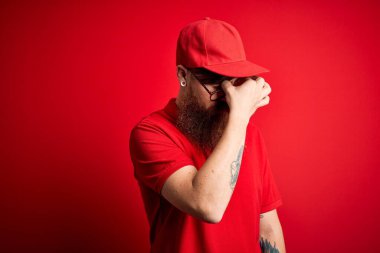 Gözlüklü ve kırmızı şapkalı genç teslimatçı izole arka planda yorgun burnu ve gözleri ovuşturup yorgun ve baş ağrısı çekiyor. Stres ve hüsran kavramı.