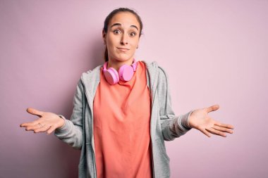 Spor yapan, spor yapan güzel bir kadın pembe arka planda kulaklık takarak müzik dinliyor ve elleri ve kolları havada, kafası karışmış bir ifade takınıyor. Şüphe kavramı.