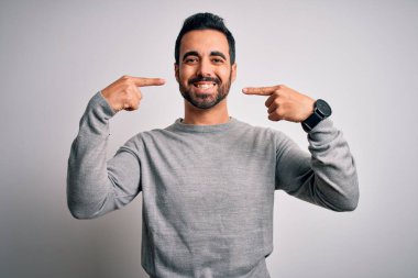 Sakallı, sıradan bir süveter giyen, beyaz arka plan üzerinde gülümseyen, parmaklarıyla ve ağzıyla işaret eden genç yakışıklı bir adam. Diş sağlığı konsepti.