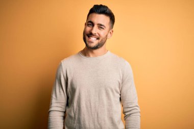 Yüzünde mutlu ve havalı bir gülümsemeyle izole edilmiş sarı arka plan üzerinde duran sıradan bir süveter giyen genç yakışıklı bir adam. Şanslı kişi..