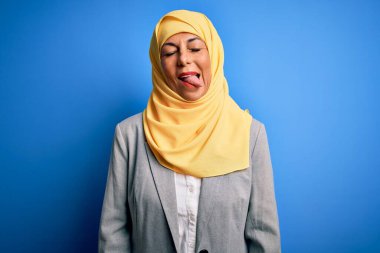 Orta yaşlı esmer iş kadını, geleneksel Müslüman tesettüre bürünmüş mavi arka planda, komik bir ifadeyle dilini dışarı çıkartıyor. Duygu kavramı.