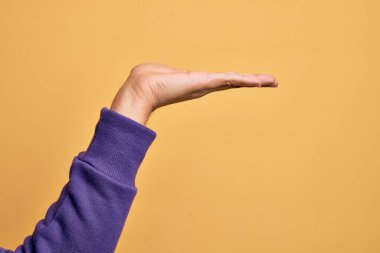 Beyaz bir gencin eli, izole edilmiş sarı arka plan üzerinde parmaklarını gösteriyor. Düz palmiye ürünü sunuyor, el hareketi yapıyor, boş kopya alanı veriyor.