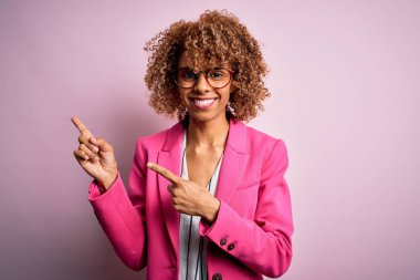 Genç Afrikalı Amerikalı iş kadını pembe arka planda duran gözlük takıyor gülümsüyor ve kameraya bakıyor iki eliyle ve parmaklarıyla yan tarafa bakıyor..