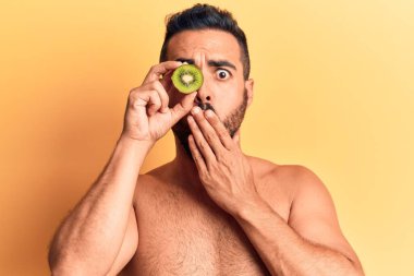 Genç İspanyol adam üstsüz duruyor, elinde kivi var. Ağzını eliyle kapatıyor. Şok olmuş ve hata yapmaktan korkuyor. şaşırmış ifade 