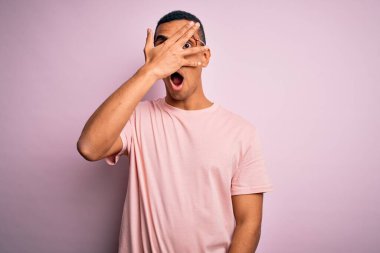 Yakışıklı Afro-Amerikalı adam, günlük tişört ve gözlük takıyor pembe arka plan üzerinde. Şok olmuş yüzünü ve gözlerini eliyle kapatıyor, utanmış ifadesiyle parmaklarından bakıyor..