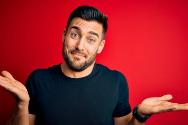 Siyah bir tişört giyen genç yakışıklı, izole edilmiş kırmızı arka plan üzerinde duruyor. Kolları ve elleri havada, kafası karışmış bir yüz ifadesi var. Şüphe kavramı.