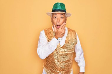 Sarı arka plan üzerinde Brezilya karnavalı kıyafeti giyen orta yaşlı, kır saçlı bir adam elleri çenesinde olan inanması güç, şaşırmış ve şaşırmış bir ifadeye sahip.