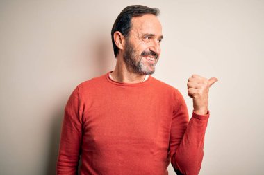 Orta yaşlı, turuncu süveter giyen, izole edilmiş beyaz arka planda dikilen mutlu yüzüyle gülümseyen ve baş parmağıyla yan tarafı gösteren orta yaşlı bir adam..