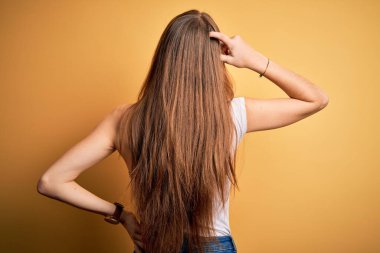 Genç, güzel, kızıl saçlı bir kadın, sarı arka planda gündelik tişörtler giyiyor. Geriye doğru, elleri kafasında şüpheyi düşünüyor.