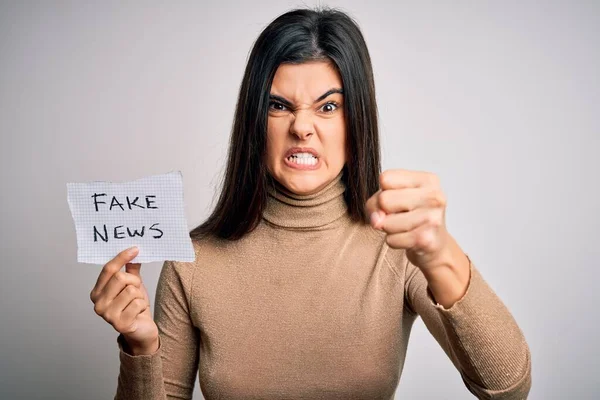 Young beautiful brunette woman holding paper with fake news message over white background annoyed and frustrated shouting with anger, crazy and yelling with raised hand, anger concept