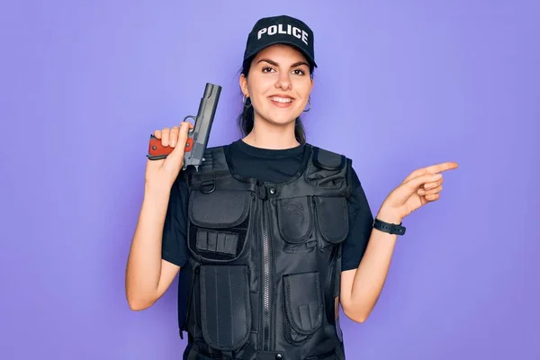 Young police woman wearing security bulletproof vest uniform and holding gun very happy pointing with hand and finger to the side