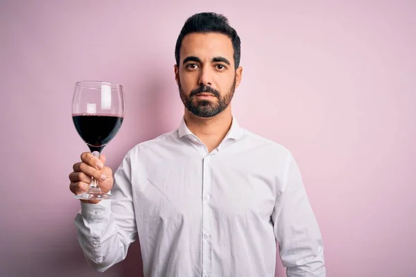 Young handsome man with beard drinking glass of red wine over isolated pink background with a confident expression on smart face thinking serious