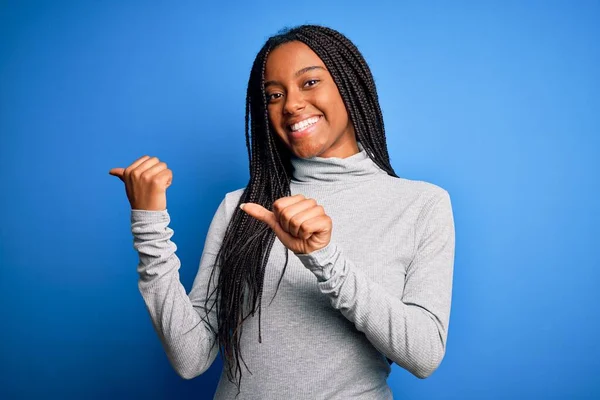 若いアフリカ系アメリカ人の女性が青い隔離された背景の上にカジュアルなタートルネックを身に着けて立っている手と親指を後ろに向け 自信を持って笑って — ストック写真