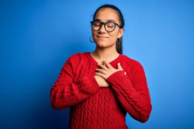 Genç, güzel Asyalı bir kadın mavi arka planda gözlük ve süveter takıyor. Elleri göğsünde, gözleri kapalı gülümsüyor ve yüzünde minnet ifadesi var. Sağlık kavramı.