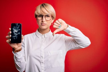 Kırık akıllı telefonu kırmızı arka planda tutan kısa saçlı genç sarışın bir kadın. Kızgın yüzlü, olumsuz işaretli, başparmakları aşağıda, reddedilme kavramından hoşlanmadığını gösteren.