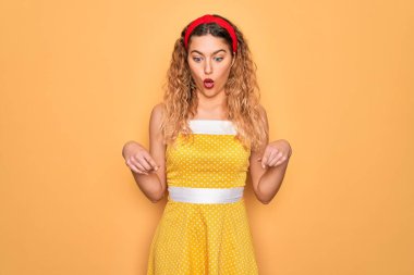 Sarı arka planda dikilip duran mavi gözlü güzel sarışın kadın parmaklarını göstererek reklamını yapıyor, şaşırmış yüzünü ve ağzını açıyor.