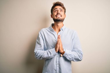 Sakallı, beyaz arka plan üzerinde çizgili gömlek giyen genç yakışıklı adam elleri bağlı yalvarıyor ve dua ediyor. Yüzünde umut ifadesi var. Çok duygusal ve endişeli. Yalvarıyorum..