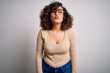 Genç ve güzel kıvırcık Arap kadın günlük tişört ve gözlük takmış beyaz arka planda kameraya bakıp havada öpüşürken güzel ve seksi görünüyor. Aşk ifadesi.