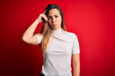 Mavi gözlü güzel sarışın kadın kırmızı arkaplan üzerine gündelik beyaz tişört giyiyor. Alnındaki sorun yüzünden endişeli ve stresli. Kriz için endişeli.