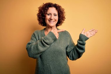 Orta yaşlı, kıvırcık saçlı, sıradan bir süveter giyen güzel bir kadın izole edilmiş sarı arka planda hayret ediyor ve kameraya gülümsüyor..