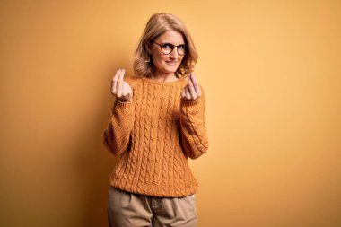 Orta yaşlı güzel sarışın kadın sarı arka planda günlük süveter ve gözlük takıyor. Elleriyle para hareketi yapıyor. Maaş ödemesi istiyor.