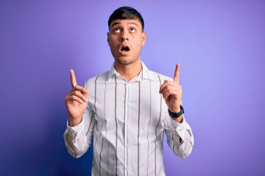 Zarif iş gömleği giyen genç yakışıklı İspanyol adam mor arka planın üzerinde şaşkın şaşkın ve şaşırmış bir şekilde yukarı bakıyor ve parmaklarıyla işaret ediyor ve kollarını kaldırıyordu..