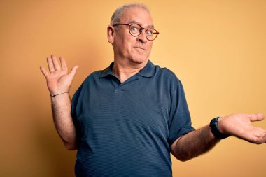 Sarı arka plan üzerine gündelik polo ve gözlük takan orta yaşlı yakışıklı ve elleri ve kolları havada, kafası karışmış bir adam. Şüphe kavramı.