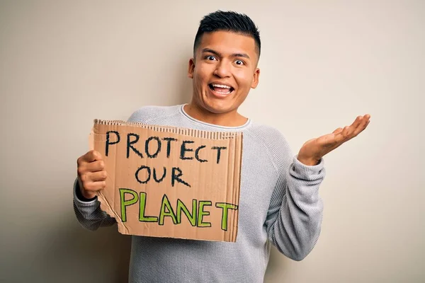 Young handsome activist latin man holding banner asking to protect our planet very happy and excited, winner expression celebrating victory screaming with big smile and raised hands