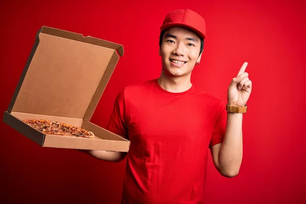 Young handsome chinese delivery man holding deliver box with delicious Italian pizza surprised with an idea or question pointing finger with happy face, number one