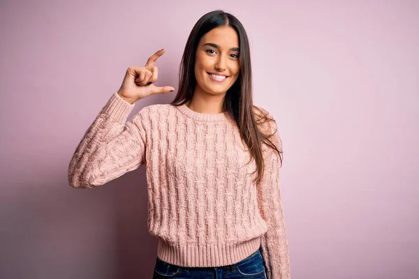 Young beautiful brunette woman wearing casual sweater over isolated pink background smiling and confident gesturing with hand doing small size sign with fingers looking and the camera. Measure concept.