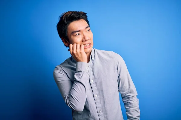 年轻英俊的中国男人穿着休闲装站在孤立的蓝色背景上 看上去很紧张 双手放在咬指甲的嘴上 焦虑问题 — 图库照片