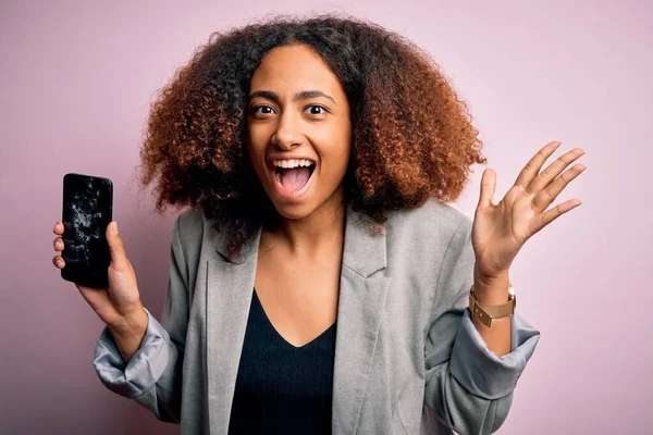 Young african american woman with afro hair holding cracked and broken smartphone screen very happy and excited, winner expression celebrating victory screaming with big smile and raised hands