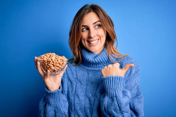 Young Beautiful Brunette Woman Holding Bowl Peanuts Blue Background Pointing — Stock Photo, Image